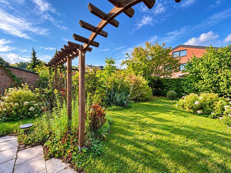 Ein gepflegter Garten mit einem Rasen und bunten Blumenbeeten. Im Hintergrund steht eine Pergola aus Holz, umgeben von verschiedenen Pflanzen und Sträuchern. Der Himmel ist blau mit einigen Wolken, und es sind Bäume in der Nähe sichtbar.