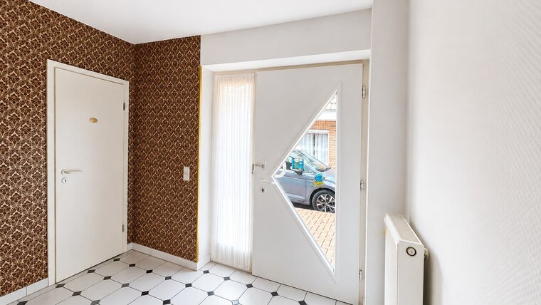Ein heller Eingangsbereich mit einem weißen, diamantförmigen Eingangstür. Die Wand ist mit brauner, gemusterter Tapete dekoriert. Rechts befindet sich ein Heizkörper, und der Boden besteht aus schwarz-weißen Fliesen. Ein Fenster in der Tür bietet einen Blick nach draußen.