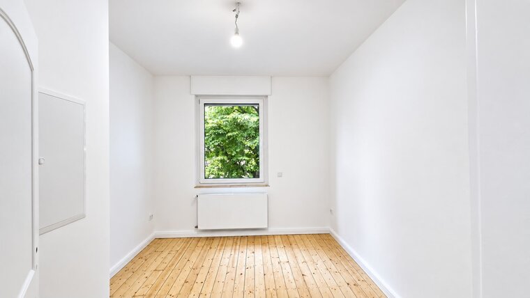 Ein leerer Raum mit weißen Wänden und einem Holzfußboden. An einem Fenster ist ein Heizkörper angebracht, durch das Fenster sind grüne Bäume sichtbar. Eine einfache Deckenleuchte sorgt für Beleuchtung. Der Raum wirkt hell und einladend.