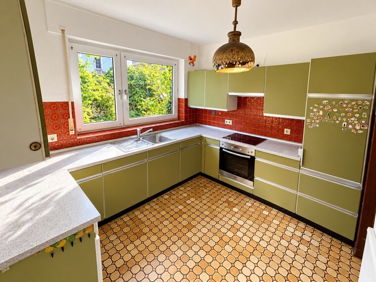 Eine helle Küche mit grünen Schränken und roter Fliesenrückwand. Ein Fenster lässt Tageslicht herein und zeigt grüne Bäume im Freien. Der Boden ist mit gelben, hexagonalen Fliesen gestaltet. Die Küche ist aufgeräumt mit einem Spülbecken und einem Herd.