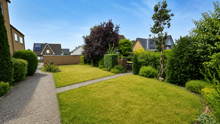 Grüner Garten mit gut gepflegtem Rasen und verschiedenen Sträuchern. Ein gepflasterter Weg führt durch den Garten. Im Hintergrund sind mehrere Wohnhäuser mit unterschiedlichen Dachformen und Pflanzen zu sehen. Der Himmel ist klar und blau.