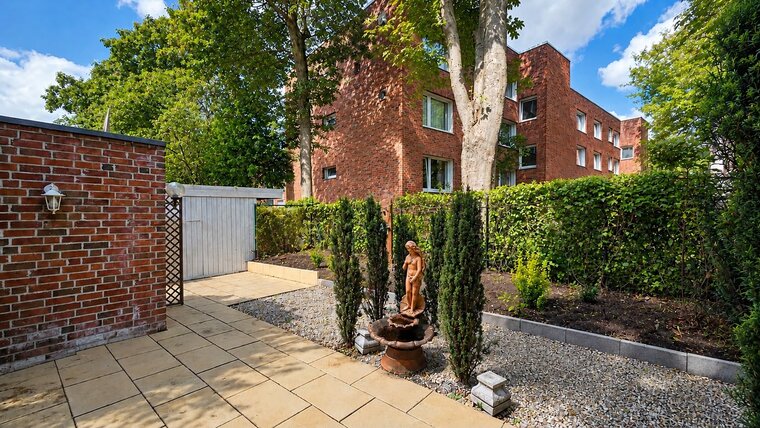 Ein hübscher Gartenbereich mit gepflastertem Weg, umgeben von Hecken und kleinen Bäumen. In der Mitte steht ein Brunnen mit einer statuarischen Figur. Im Hintergrund sind Ziegelgebäude und ein helles, blaues Himmel zu sehen.