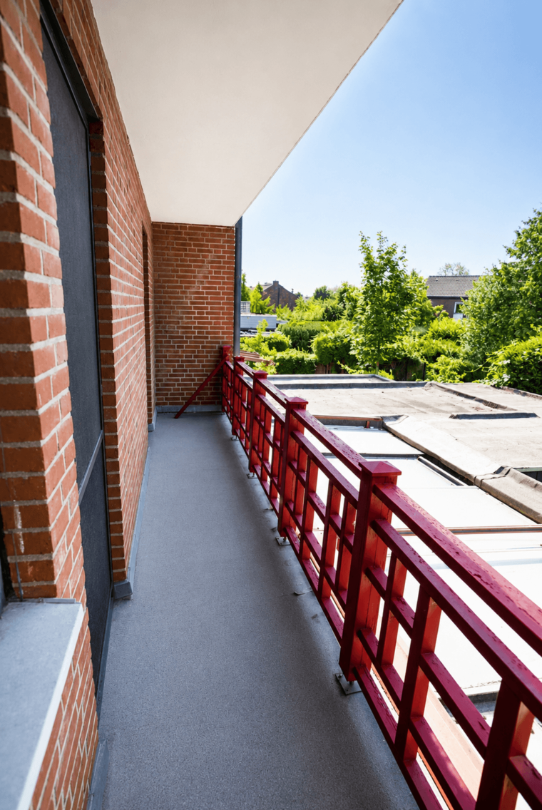 Ein Balkon mit roter Brüstung und einem grauen Bodenbelag. Im Hintergrund sind grüne Bäume und Gebäude sichtbar. Der Himmel ist klar und sonnig. Es gibt eine abgeschlossene Wand und einen Zugang zum Haus.