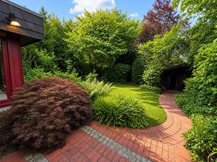 Ein gepflegter Garten mit üppigem Grün, bunten Pflanzen und einem geschwungenen roten Gehweg. Im Hintergrund befindet sich eine kleine Holzhütte, umgeben von dichten Sträuchern und Bäumen, die eine ruhige Atmosphäre schaffen. Der Himmel ist blau mit einigen Wolken.
