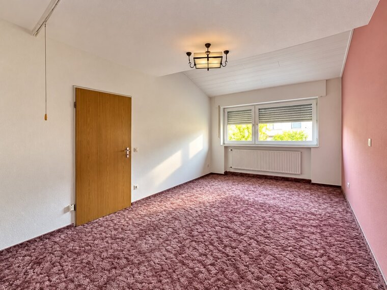 Ein leerer Raum mit einem braunen Teppichboden. An der wandseitigen Wand befindet sich ein großes Fenster mit Rolläden und ein Heizkörper. Eine Holzinnentür führt in einen angrenzenden Raum. Die Wände sind in einem hellen Farbton gestrichen, eine Wand ist pink. Ein einfaches Deckenlicht ist vorhanden.