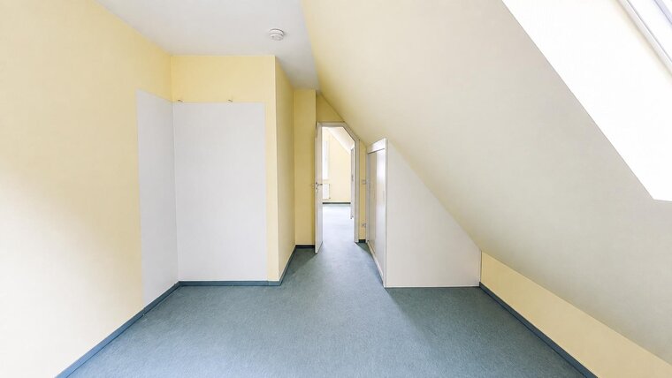 Ein heller, leerer Raum mit gelben Wänden und einem blauen Bodenbelag. Rechts befindet sich eine offene Tür zu einem weiteren Raum, während an der linken Wand ein geschlossener Schrank steht. Der Raum hat eine schräge Decke.