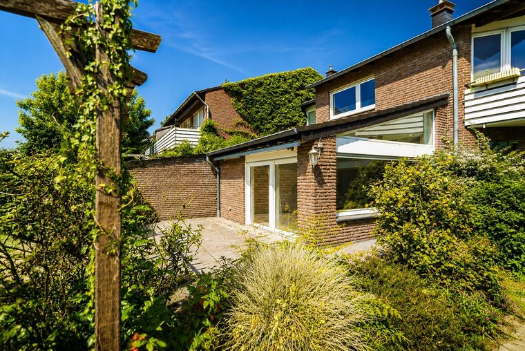 Eine moderne, brickverkleidete Hausfassade mit großen Fenstern, umgeben von üppigem Grün und einem gepflasterten Außenbereich. Der Himmel ist blau und klar, was auf einen sonnigen Tag hindeutet. Ein Holzgestell mit Pflanzen ist im Vordergrund sichtbar.