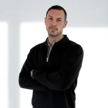 Ein Mann steht mit verschränkten Armen und einem neutralen Ausdruck. Er trägt einen schwarzen Pullover und hat kurze Haare. Der Hintergrund ist hell und minimalistisch, was den Fokus auf die Person lenkt.