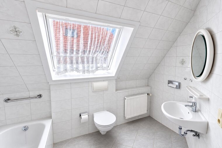Das Bild zeigt ein modernes Badezimmer mit weißen Fliesen und einer schrägen Decke. Es enthält eine Badewanne, ein wandhängendes WC, ein Waschbecken mit Spiegel und einen Heizkörper. Ein Fenster lässt Tageslicht herein, während ein weißer Vorhang das Licht filtert.