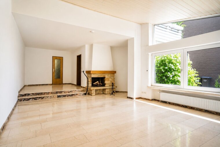 Ein heller, offener Raum mit glänzendem Marmorboden. An einer Wand befindet sich ein Kamin aus Stein mit Holz. Links gibt es eine Treppe und zwei Türen, während große Fenster den Raum mit Licht erfüllen und einen Blick ins Grüne bieten.
