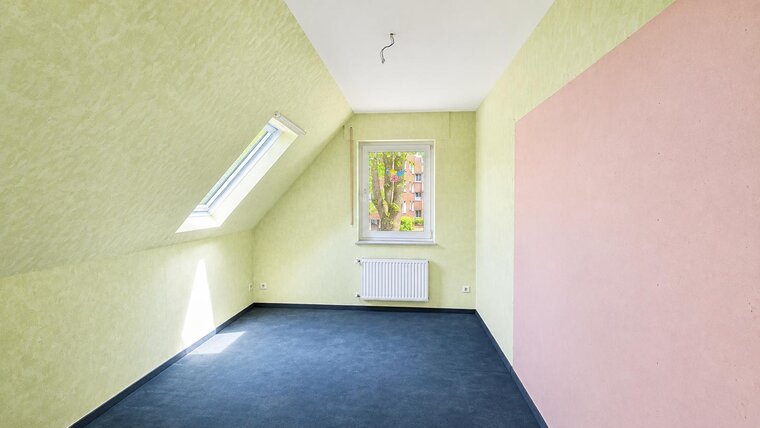 Das Bild zeigt einen leeren Raum mit schrägen Wänden und grünem Putz. Ein Fenster lässt Tageslicht hinein, und eine Heizung ist an der Wand sichtbar. Der Fußboden ist dunkelblau, während eine Wand im Raum rosa gestrichen ist.