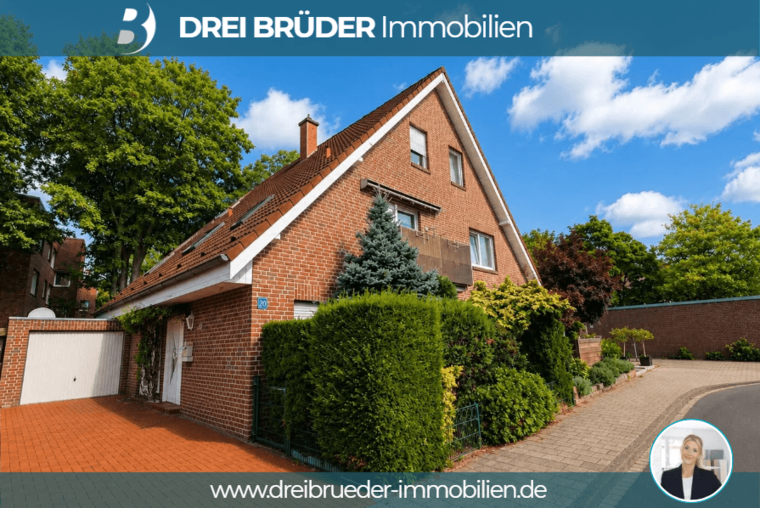 Ein modernes, zweigeschossiges Einfamilienhaus aus Ziegelstein befindet sich auf einem gepflegten Grundstück, umgeben von grünen Büschen und Bäumen. Der Eingangsbereich ist mit einem kleinen überdachten Vorbau versehen. Die Fassade zeigt große Fenster und ein Satteldach.