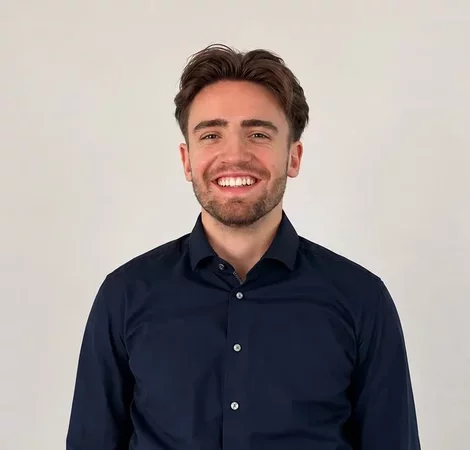 Ein junger Mann mit kurzen, braunen Haaren und einem gepflegten Bart trägt ein dunkelblaues Hemd. Er steht vor einem neutralen, hellen Hintergrund und lächelt freundlich in die Kamera.