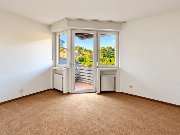 Ein heller Raum mit Teppichboden, der einen Balkon und große Fenster zeigt. Durch die Fenster ist eine grüne Landschaft mit Bäumen und Häusern sichtbar. Der Raum hat eine neutrale Farbe und ist leer, mit einer Heizung an der Wand.