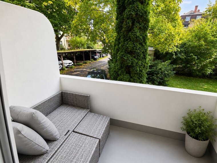 Ein moderner Balkon mit einem grauen Sitzbereich aus Rattan und zwei Kissen. Im Hintergrund sind grüne Bäume und ein Garten sichtbar. Die Atmosphäre ist ruhig und einladend, ideal zum Entspannen im Freien.