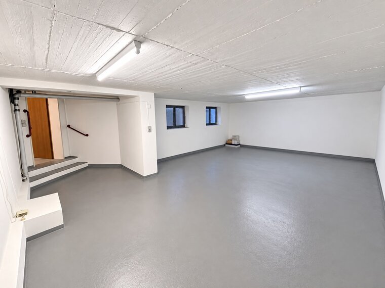 Ein heller, weißer Raum mit grauem Bodenbelag. An der Wand befindet sich eine Treppe, die zu einer Tür führt. Links sind Fenster, durch die Tageslicht hereinfällt. Der Raum ist leer und aufgeräumt.