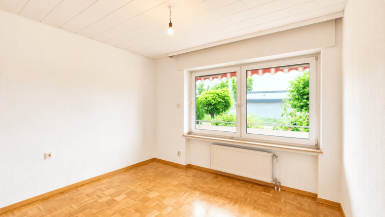 Ein heller, leerer Raum mit einem Holzfußboden. An einer Wand befindet sich ein großes Fenster, das einen Blick auf grüne Bäume und Sträucher bietet. Die Wände sind einfach und weiß, mit einer einzigen Glühbirne an der Decke.