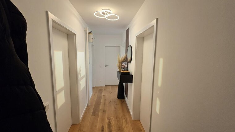 Ein heller Flur mit zwei geschlossenen Türen auf der linken und rechten Seite. An der Wand hängt ein runder Spiegel. Eine schwarze Konsole steht an der Wand, dekoriert mit Trockenblumen und einer Zeitschrift. Der Boden ist aus hellem Holz.