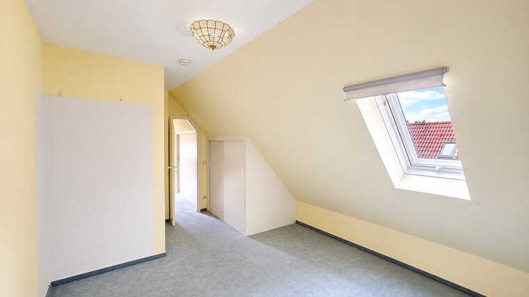 Ein heller Raum mit schrägen Wänden und einem Dachfenster. Die Wände sind in Gelb gestrichen, und der Boden ist mit grauem Teppich ausgestattet. Links steht ein kleiner weißer Schrank, und im Hintergrund ist ein Flur zu sehen.