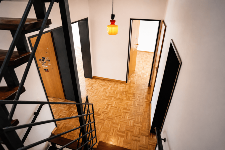 Ein Blick auf eine Treppe aus Holz mit metallischem Geländer, die in einen hellen Flur führt. Der Boden ist aus Parkett. An der Decke hängt eine rote Lampe. Links und rechts führen Türen in verschiedene Zimmer.