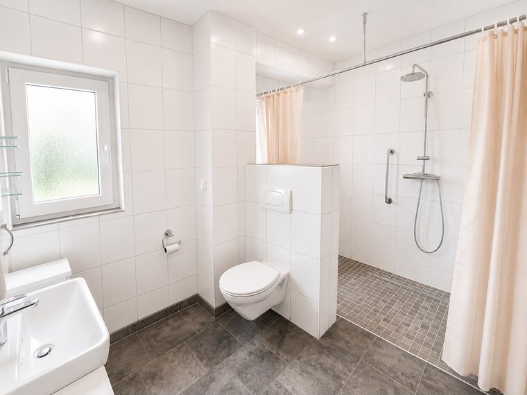 Das Bild zeigt ein modernes Badezimmer mit weißen Fliesen. Es enthält eine Dusche mit Glasabtrennung, ein Waschbecken und eine Toilette. Ein Fenster lässt Tageslicht herein, und die Dusche hat eine Handbrause sowie eine Halterung. Die Wände sind hell und die Einrichtung minimalistisch.