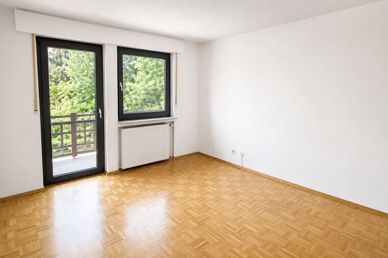 Das Bild zeigt einen leeren Raum mit Parkettboden. Auf der linken Seite befinden sich zwei große Fenster mit Blick auf grüne Bäume. An der Wand unter dem Fenster ist ein Heizkörper angebracht. Die Wände sind weiß, und der Raum wirkt hell und einladend.