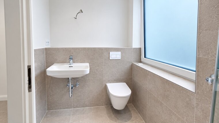 Das Bild zeigt ein modernes Badezimmer mit einem weißen Waschbecken und einer hängenden Toilette. Die Wände sind in neutralen, hellen Farbtönen gefliest. Ein großes Fenster mit frosted Glas bringt Tageslicht in den Raum. Es gibt einen Handtuchhalter an der Wand.