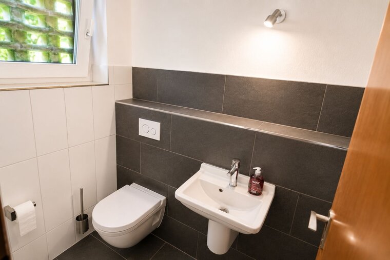 Das Bild zeigt ein modernes Badezimmer mit einer Toilette und einem Waschbecken. Die Wände sind teilweise gefliest, und es gibt einen Wandspiegel sowie einen Seifenspender auf der Ablage. Ein Fenster sorgt für natürliches Licht.