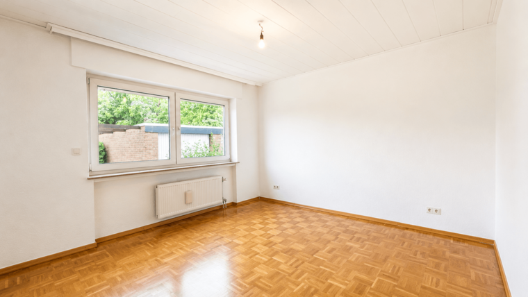 Der Raum ist leer und hat hell gestrichene Wände. Ein großes Fenster lässt viel Tageslicht herein, während ein Heizkörper an der Wand angebracht ist. Der Boden besteht aus einem hellen, geflammten Parkett. Hinter dem Fenster sind grüne Pflanzen zu sehen.