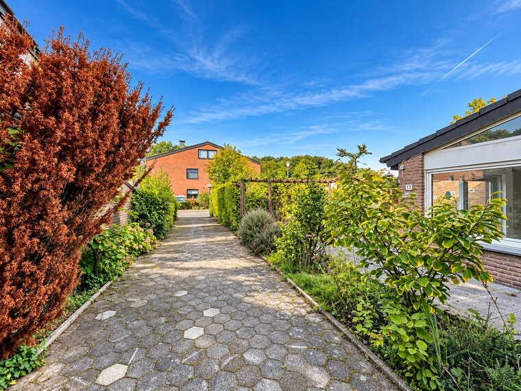 Ein gepflasterter Weg führt durch einen Garten mit verschiedenen Pflanzen und Sträuchern. Auf der linken Seite steht ein hellbrauner Busch mit roten Blüten. Im Hintergrund sind Wohnhäuser und ein blauer Himmel mit einigen Wolken zu sehen.