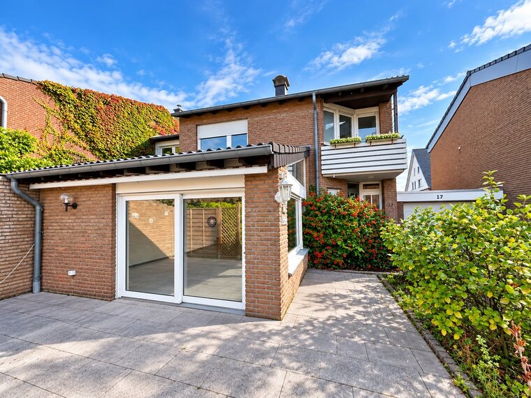 Das Bild zeigt ein modernes Haus mit roten Ziegeln und einer modernen Frontveranda. Große Fenster und eine Terrassentür bieten Zugang zu einem Innenraum. Vor dem Haus befinden sich Grünpflanzen und bunte Blumen. Der Himmel ist blau mit einigen Wolken.